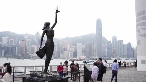 Avenue-of-stars-in-Tsim-Sha-Tsui,-Hong-Kong,-black-statue-of-a-woman-and-people-walking,-on-the-horizon-the-sea-and-the-skyline-of-the-city