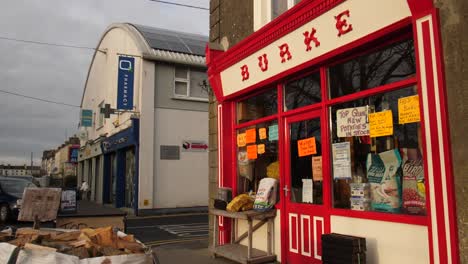 Burke-Laden-In-Gort,-Malerische-Rote-Fassade,-Straße-Mit-Anderen-Geschäften-Im-Hintergrund-An-Einem-Bewölkten-Tag