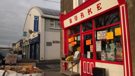 Tienda-Burke-En-Gort,-Pintoresca-Fachada-Roja,-Calle-Con-Otros-Negocios-En-El-Fondo-En-Un-Día-Nublado