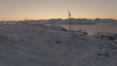 Wind-turbines-on-ridge-in-Arctic-winter-landscape-at-sunset,-drone-slow-motion