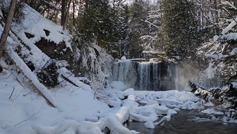 Slow-Pan-Right-View-Across-Snow-Covered-Woodland-View-Of-Hogs-Falls-With-Water-Cascading-And-Flowing-Through-Stream