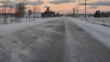 POV-shot-driving-down-a-rural-highway-after-a-snow-storm-with-snow-blowing-across-the-road-at-sunset-on-with-scatter-clouds-in-an-orange-sky