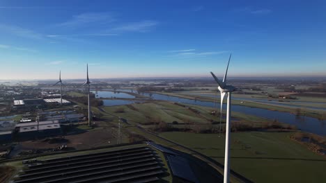 Wind-mill-turbines,-water-treatment,-solar-panels-and-bio-energy-facility-in-The-Netherlands-part-of-sustainable-industry-in-Dutch-flat-river-landscape-against-blue-sky
