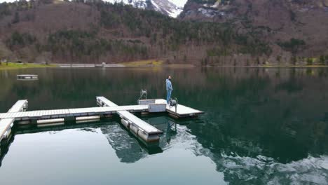 Aerial-flying-around-a-young-man-standing-on-a-lake-pier-in-the-Alps,-Switzerland
