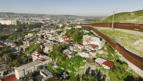 Vista-Aérea-Siguiendo-El-Muro,-Sobre-Una-Zona-Pobre,-Hacia-La-Estación-Fronteriza-En-El-Soleado-San-Ysidro,-Tijuana,-México