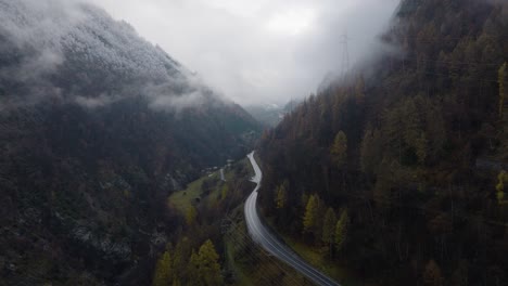 Stunning-backwards-aerial-drone-shot-in-Swiss-Alpine-cloud-covered-valley-with-mountain-road-green-trees-and-snow-covered-forest-on-cloudy-winter-afternoon-in-4K