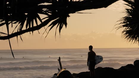 Surfer-waiting-to-enter-the-ocean-at-sunrise-at-Burleigh-Heads,-Gold-Coast,-Queensland,-Australia