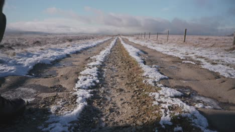 Two-people-walking-past-the-camera,-hiking-away-along-a-long,-snowy,-frozen-trail-through-open-moorland-on-a-beautiful-winter-day