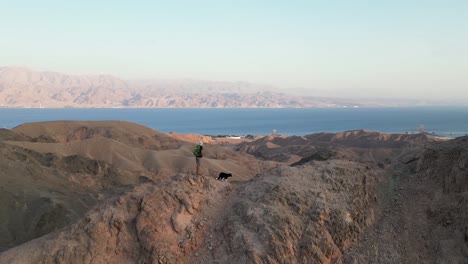 A-view-of-a-man-hiking-with-his-dog-in-the-desert-mountains,-with-a-sea-view-captured-by-a-drone
