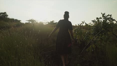 The-girl-walks-through-the-field-in-a-black-dress,-her-hand-gently-touches-the-tops-high-green-grass,-the-sun-shines-as-she-walks-through-the-sun-flare