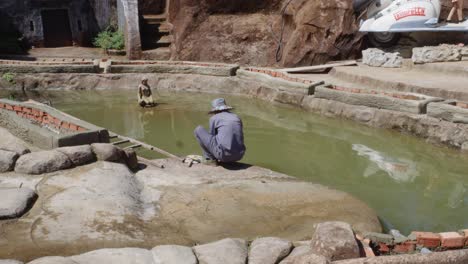 Museum-worker-crouches-by-a-pond-in-Clay-Tunnel-in-Da-Lat,-Vietnam,-washing-his-sculpting-tools