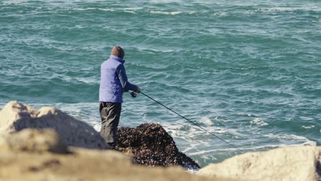 Fisherman-winding-fishing-rod-reel,-ocean-waves-in-background,-sunny-day
