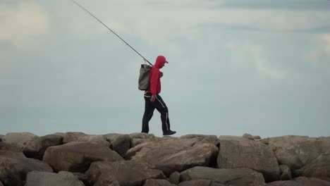Tracking-shot-of-fisherman-with-rod-walking-on-rocky-jetty-near-sea-for-fishing---slow-motion