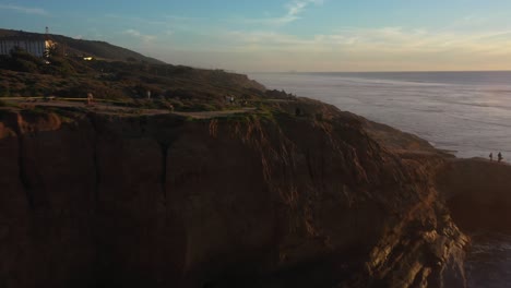 Drone-shot-follows-the-rugged-coastline-as-the-drone-flies-low-and-close-to-the-rocky-terrain,-nearly-hitting-a-massive-cliff-and-suddenly-revealing-the-entire-view-of-the-glowing-coast-ahead