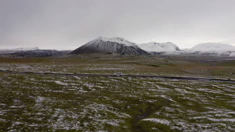 Aerial:-Side-follow-of-one-white-car-driving-on-a-remote-mountain-road-in-Iceland-highlands-partially-covered-with-snow