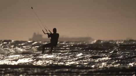 Medium-shot-of-a-silhouetted-person-kitesurfing-on-the-rough-ocean-with-a-ship-in-the-background-near-sunset