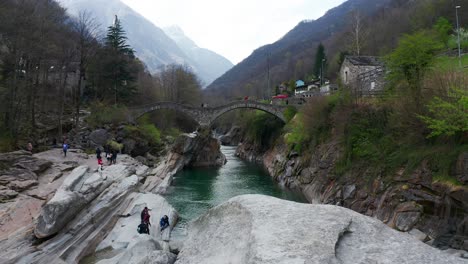 Aerial-View-Of-Banks-Of-Verzasca-River-And-Ponti-Dei-Salti-Bridge,-Lavertezzo,-Ticino,-Switzerland-With-Tourists-Exploring