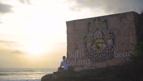 Ein-Mann-Sitzt-Am-Strandschild-Von-Batu-Bolong,-Während-Sich-Die-Menschen-Entspannen,-Während-Die-Sonne-Untergeht