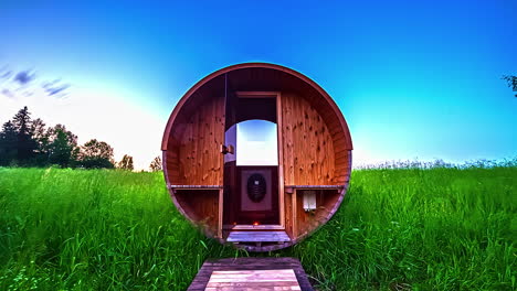 Timelapse-of-a-round-shaped-wooden-cabin-on-a-green-meadow-under-blue-sky-and-moving-clouds-at-dusk