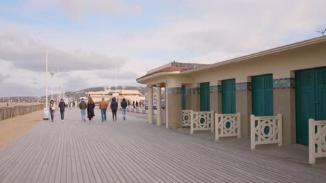 Gente-Caminando-En-El-Paseo-Marítimo-De-Les-Planches-A-Lo-Largo-De-Las-Cabañas-De-Baño-En-Un-Día-Frío-En-La-Playa-De-Deauville,-Normandía,-Francia