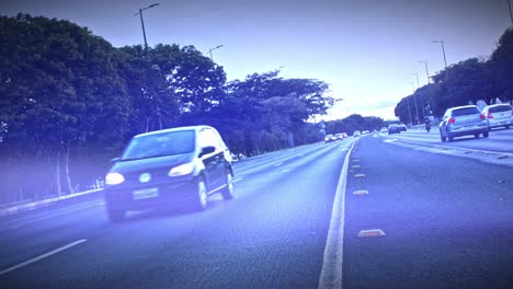 Clip-from-the-center-lane-as-cars-travel-along-the-road-at-dusk-just-after-light-rain