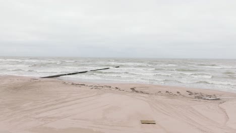 Establishing-aerial-view-of-Baltic-sea-coast-on-a-overcast-day,-old-wooden-pier,-white-sand-beach,-large-storm-waves-crushing-against-the-coast,-climate-changes,-wide-drone-shot-moving-forward-low
