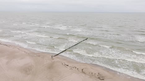 Establishing-aerial-view-of-Baltic-sea-coast-on-a-overcast-day,-old-wooden-pier,-white-sand-beach,-large-storm-waves-crushing-against-coast,-climate-changes,-wide-drone-shot-moving-forward-tilt-down