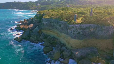 Aerial-backwards-shot-of-green-woodland-landscape-on-hill-with-rocky-cliff-and-coastline-during-sunset-time---Strong-waves-of-Caribbean-Sean-at-CABO-FRANCES-VIEJO