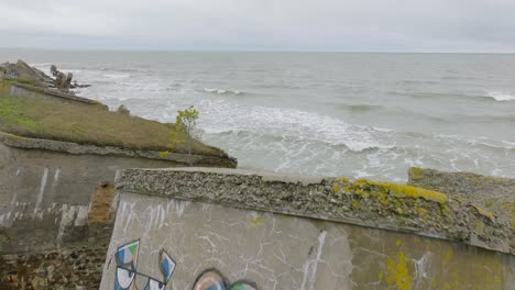 Aerial-establishing-view-of-abandoned-seaside-fortification-buildings-at-Karosta-Northern-Forts,-Baltic-sea-coast-,-large-waves,-overcast-day,-slow-motion,-drone-shot-moving-forward