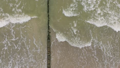 Aerial-birdseye-view-of-Baltic-sea-coast-on-a-overcast-day,-old-wooden-pier,-white-sand-beach,-large-storm-waves-crushing-against-the-coast,-climate-changes,-wide-drone-shot-moving-forward