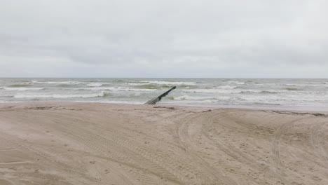 Establishing-aerial-view-of-Baltic-sea-coast-on-a-overcast-day,-old-wooden-pier,-white-sand-beach,-large-storm-waves-crushing-against-the-coast,-climate-changes,-wide-drone-shot-moving-forward-low