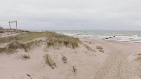 Establishing-aerial-view-of-Baltic-sea-coast-on-a-overcast-day,-old-wooden-pier,-white-sand-beach,-large-storm-waves-crushing-against-the-coast,-climate-changes,-ascending-drone-shot-moving-forward