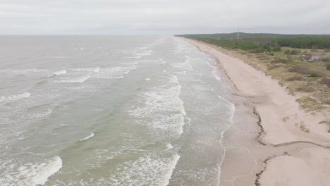 Establishing-aerial-view-of-Baltic-sea-coast-on-a-overcast-day,-old-wooden-pier,-white-sand-beach,-large-storm-waves-crushing-against-the-coast,-climate-changes,-wide-drone-shot-moving-forward-high