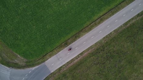 Slowly-turning-aerial-drone-shot-of-motorcyclist-driving-down-a-road-between-grass-field