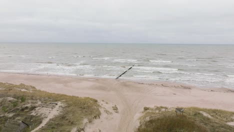Establishing-aerial-view-of-Baltic-sea-coast-on-a-overcast-day,-old-wooden-pier,-white-sand-beach,-large-storm-waves-crushing-against-the-coast,-climate-changes,-wide-drone-shot-moving-forward