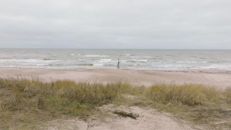 Establishing-aerial-view-of-Baltic-sea-coast-on-a-overcast-day,-frame-Jurmalciems,-old-wooden-pier,-white-sand-beach,-large-storm-waves-crushing-against-the-coast,-wide-drone-shot-moving-backward