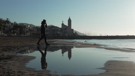 Against-backdrop-of-coastal-town-where-church-is-reflected-in-water,-man-walks-in-frame-and-stops-before-sea-as-waves-gently-washes-ashore