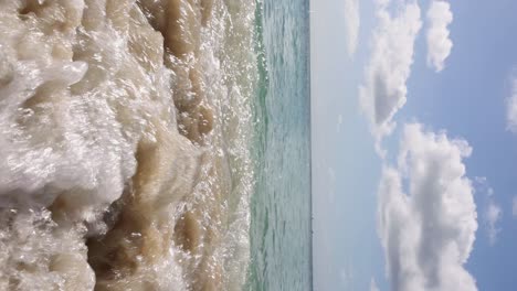 Low-angle-view-of-small-waves-on-beach-with-clear-blue-water-and-fluffy-clouds-in-sky