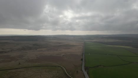Aerial-drone-footage-revealing-a-long,-straight-road-through-open-fields-and-moorland-with-a-dramatic,-cloudy-sky