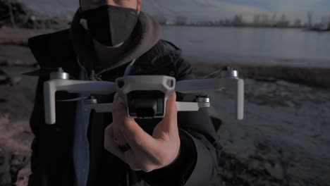 Mann-Mit-Stoffmaske,-Der-Einen-Kleinen-Drohnen-UAV-Quadcopter-In-Der-Hand-Hält-Und-Zeigt,-Mit-Grasstrand,-Sandwasserfluss-Im-Hintergrund