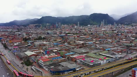 Panoramic-wide-shot-of-Bogota's-skyline-and-mountains,-slowly-revealing-an-important-intersection-with-an-elevated-round-point-allowing-for-red-articulated-buses-to-flow-from-a-street-to-a-main-avenue