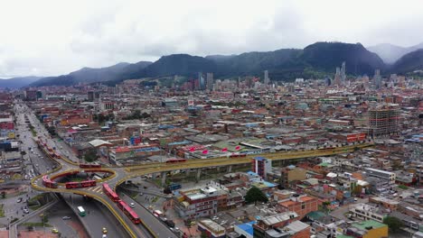 Toma-Aérea-De-Seguimiento-De-Carros-Del-Horizonte-De-Bogotá,-Comenzando-En-Una-Intersección-Importante-Con-Un-Punto-Redondo-Elevado-Que-Permite-Que-Los-Autobuses-Articulados-Rojos-Fluyan-De-Una-Calle-A-Una-Avenida-Principal