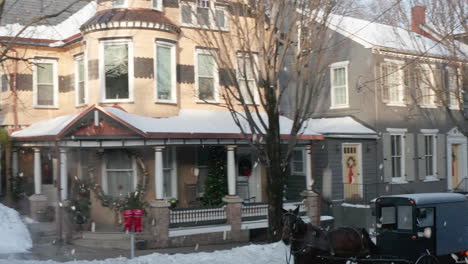 Amish-horse-and-buggy-along-Lititz,-PA-street-with-colonial-homes-decorated-for-Christmas-winter-holidays,-snow-falling