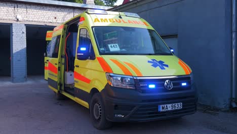 The-yellow-paramedic-ambulance-vehicle-van-parked-outside-the-garage-with-blue-flashing-warning-lights,-sunny-day,-handheld-medium-shot
