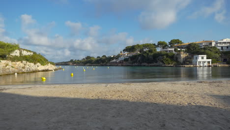 Malerische-Aussicht-Bei-Sonnenaufgang-Auf-Den-Strand-An-Der-Bucht-Cala-Santandria-Auf-Menorca-Mit-Gelben-Bootsmarkierungen,-Blauem-Meer-Und-Umliegenden-Felsen