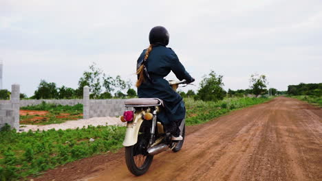 Woman-with-long-red-hair-ponytail-riding-motorbike-wearing-helmet-through-dirt-road,-back-view