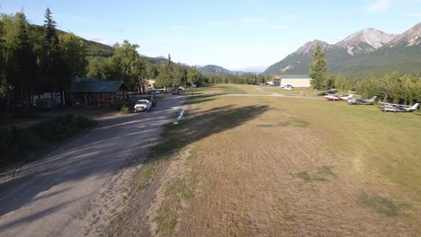 Imágenes-De-Drones-Volando-Lentamente-Sobre-Una-Pista-De-Césped-Privada-Con-Vehículos-Estacionados-A-Lo-Largo-De-Los-Edificios-En-Las-Montañas-Talkeetna