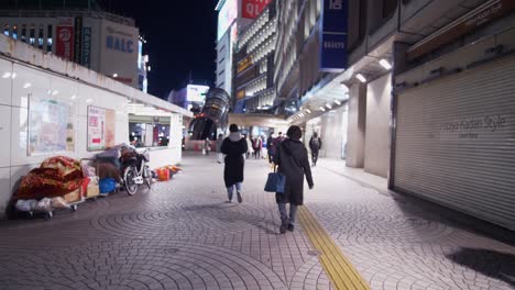 Weitwinkelaufnahme-Hinter-Einer-Frau,-Die-Geht,-Und-Anderen-Menschen,-Die-Masken-Tragen,-Nachts-Vor-Einem-Ruhigen-Bahnhof-In-Shinjuku,-Vorbei-An-Einem-Obdachlosen,-Der-Draußen-Schläft-Und-Geschäfte-Geschlossen-Hat