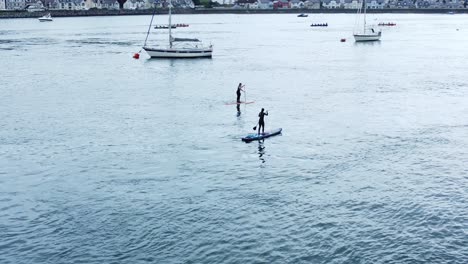 Girls-standing-balancing-paddle-boarding-floating-down-river-near-sailboat-aerial-tracking-view