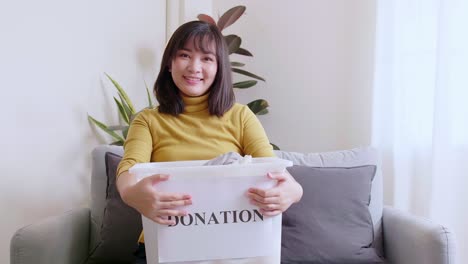Portrait-of-young-Asian-woman-looking-at-camera-holding-box-with-clothes-for-donation